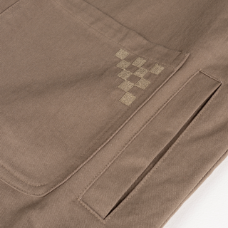 Close-up of a tan cotton work jacket with a chest pocket and two patch pockets featuring checkerboard motif embroidery.