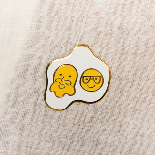 Sunny Side Up Pin of the Month: Gold-trimmed pin of a fried egg with two sunny-side-up yolks smiling with a mustache and glasses.