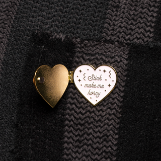 Heart-shaped locket pin. One gold heart is hinged open, revealing "Stink make me horny" text on the white enamel heart.